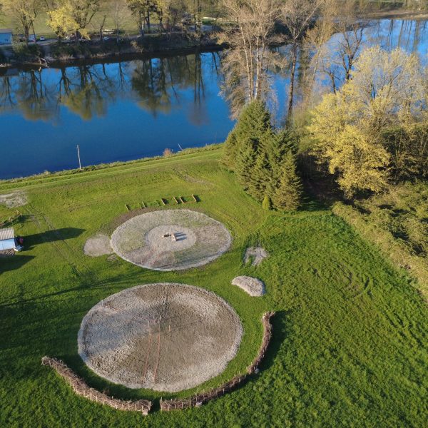 Aerial view of grassy land by a river, featuring two large circular structures outlined with stones, surrounded by trees and a few small buildings.